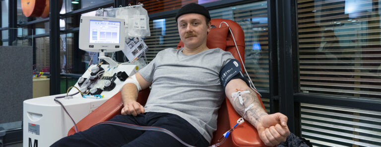 Man donating platelets. An apheresis device can be seen next to it, and a smiley drop from the Blood Service in the background.