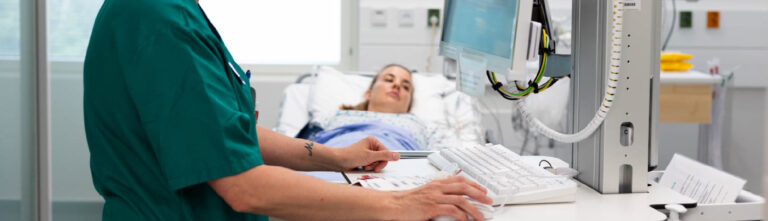 Potilas makaa sairaalasängyllä, etualalla sairaanhoitaja katsoo tietoja tietokoneelta.