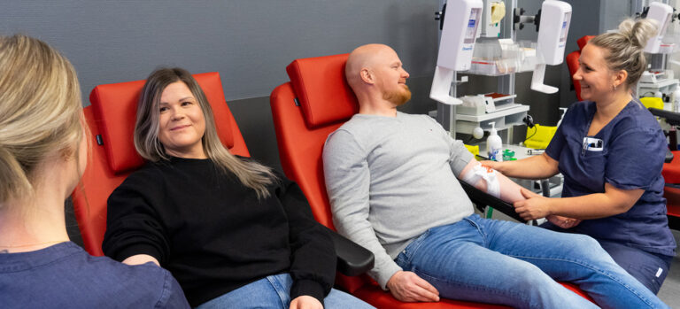 Two blood donors sit smiling while donating blood. Two nurses are sitting next to them.