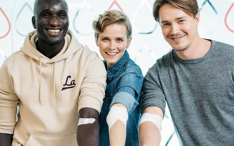 Kolme verenluovuttajaa ojentaa käsiään ja hymyilee kameralle.