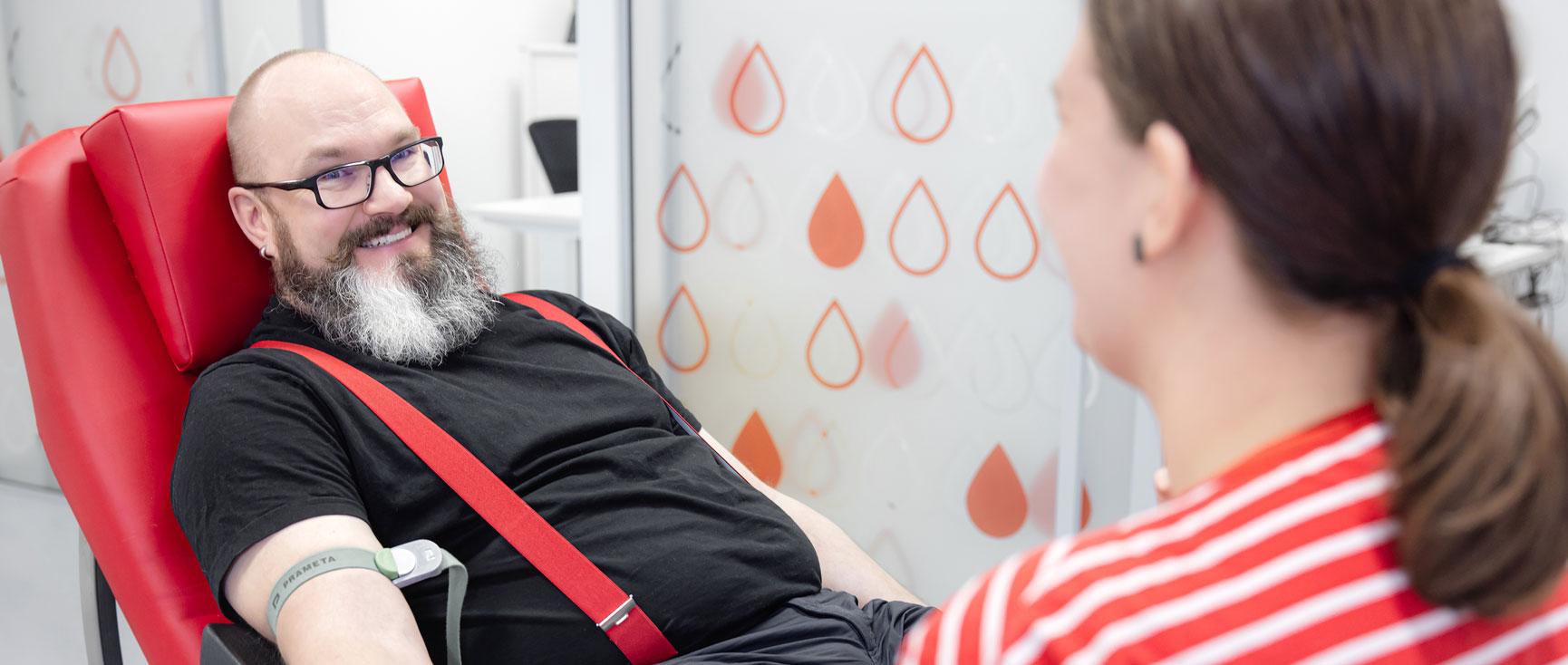 Blooddonor is looking at the camera, a nurse is in front in the picture.